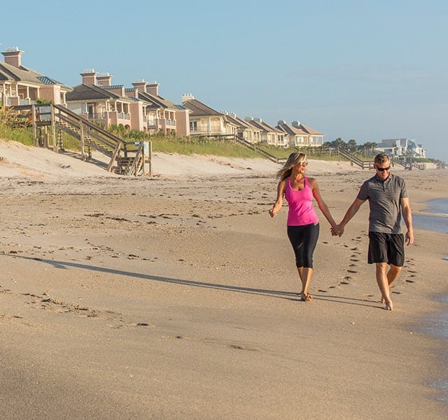 Couple walk along the beach, holding hands, in front of the Orchid Island Condos