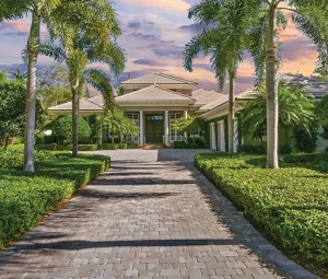 Palm-lined stone driveway leading to a large tropical-style home at sunset.