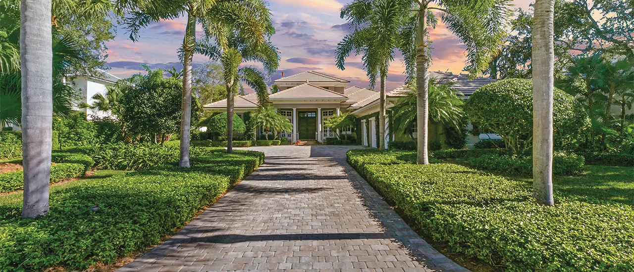 Palm-lined stone driveway leading to a large tropical-style home at sunset.