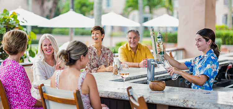 Adults socializing at an outdoor bar while a server pours a drink.