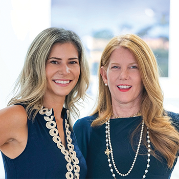 Two women standing side by side indoors, smiling at the camera.