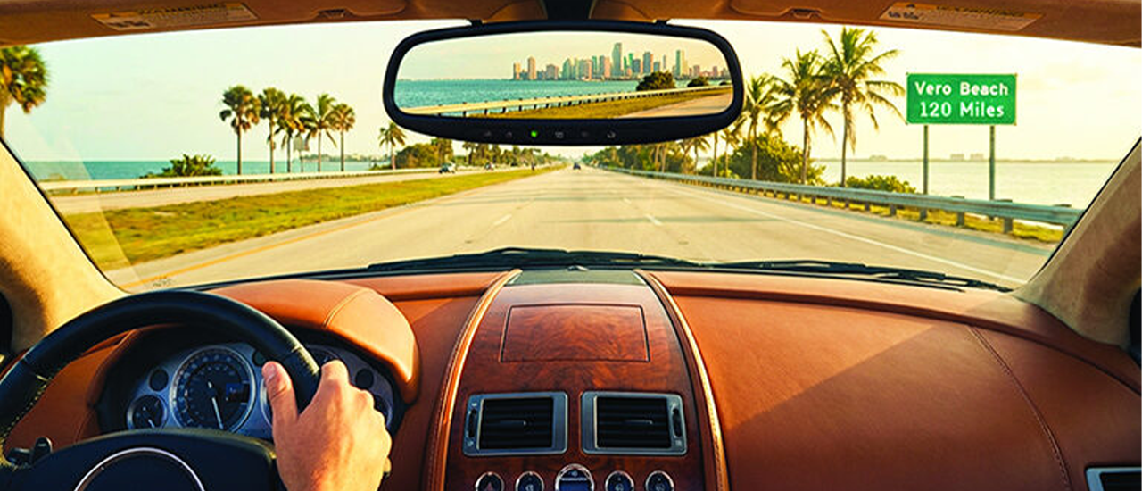 View from inside a convertible driving along a palm-lined coastal highway with ocean and road sign ahead
