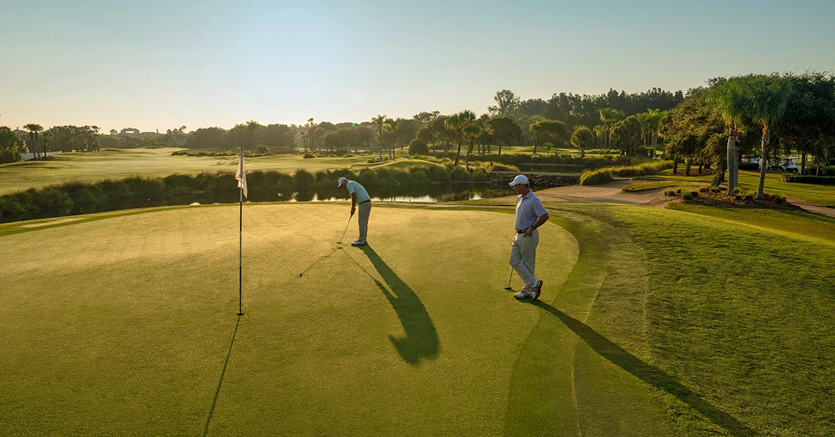 Two golfers stand on a putting green at sunset.