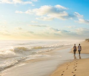 Two people walk along a sandy beach beside ocean waves under a bright blue morning sky.