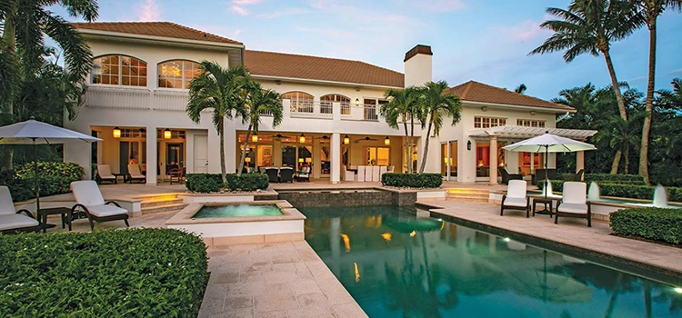 Twilight exterior of a large coastal residence with a rectangular pool, raised spa, stone patio, palm trees, lounge chairs, umbrellas, and warm interior lighting.