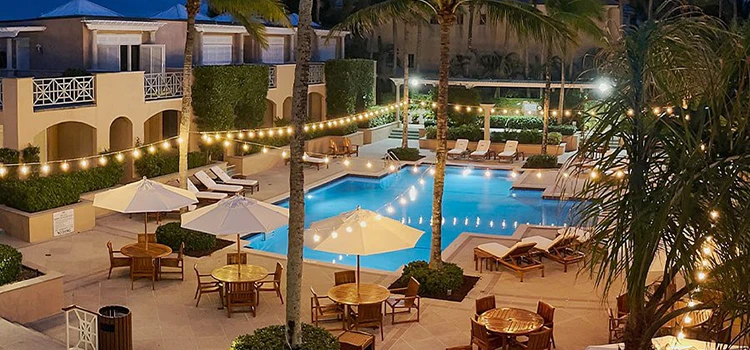 Evening pool courtyard with palm trees, lounge chairs, umbrellas, and glowing string lights.