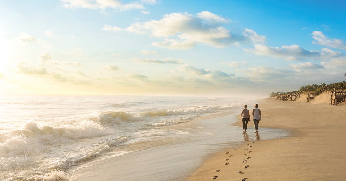 Two people walk along a sandy beach beside ocean waves under a bright blue morning sky.