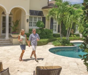 A couple walks hand-in-hand across the elegant stone patio of their luxury Mediterranean-style home, featuring arched columns, palm trees, and a sparkling sw...