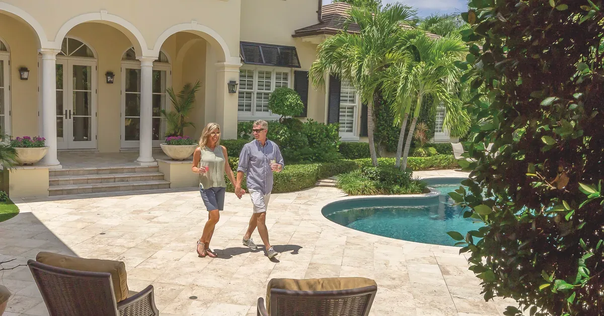 A couple walks hand-in-hand across the elegant stone patio of their luxury Mediterranean-style home, featuring arched columns, palm trees, and a sparkling sw...
