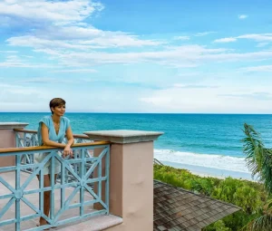 Woman enjoys ocean views from the balcony of a luxury beachfront home with turquoise waters and palm trees in tropical paradise.