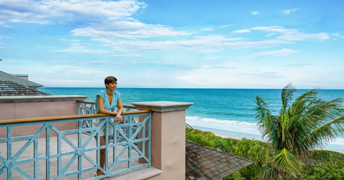 Woman enjoys ocean views from the balcony of a luxury beachfront home with turquoise waters and palm trees in tropical paradise.