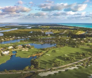 Aerial view of a luxury coastal golf community with pristine fairways, serene lakes, elegant homes, and turquoise ocean waters stretching to the horizon.